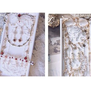 May include: Two jewelry sets displayed on white rectangular trays. The left set, labeled Full Set, Red, features gold-tone chains, red teardrop stones, and cross charms. The right set, labeled Full Set, Light-pink, includes gold-tone chains, butterfly charms, and light pink accents.