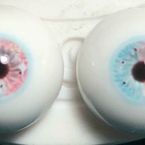 May include: Two realistic-looking glass eyes with a blue and pink iris and a brown pupil. The eyes are set on a white surface.