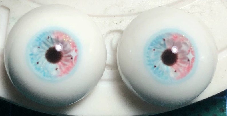 May include: Two realistic-looking glass eyes with a blue and pink iris and a brown pupil. The eyes are set on a white surface.