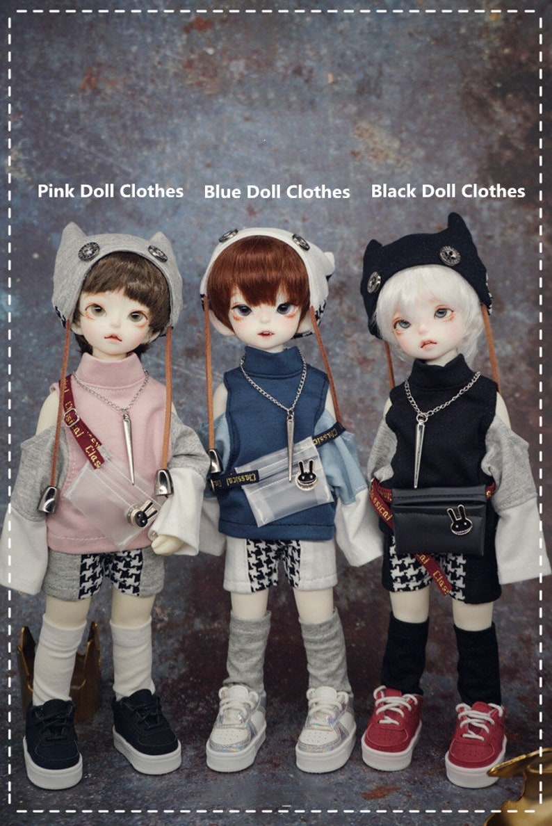 May include: Three dolls wearing different outfits. The doll on the left is wearing a pink shirt, grey pants, and a grey cat-eared hat. The doll in the middle is wearing a blue shirt, blue pants, and a grey cat-eared hat. The doll on the right is wearing a black shirt, black pants, and a black cat-eared hat. All three dolls are wearing white socks and sneakers.