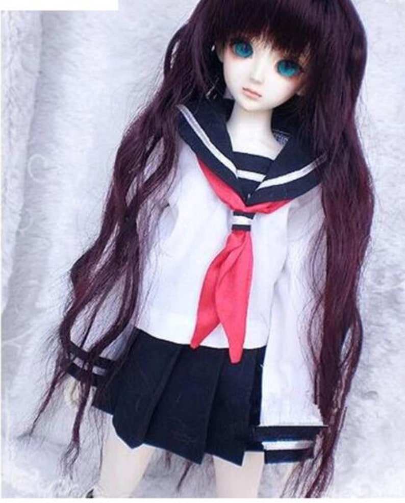May include: A doll wearing a white sailor uniform with a red tie and a black pleated skirt. The doll has long, dark brown hair and blue eyes.