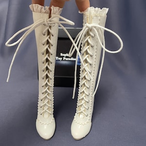 Pode incluir: Um par de botas brancas com atacadores e cano alto. As botas t&ecirc;m bico redondo e uma babado decorativo no topo. Os atacadores s&atilde;o brancos e cruzam-se na frente. As botas s&atilde;o feitas de um material liso, possivelmente semelhante a couro.