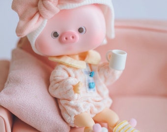 May include: A pink pig doll wearing a white and pink hat with a bow, a pink and yellow sweater, and yellow and white striped socks. The doll is sitting on a pink couch and holding a white cup.