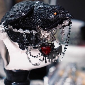 K&ouml;nnte beinhalten: Eine schwarze Rabenfigur mit einem silbernen Schmetterlingsanh&auml;nger, einem roten Herzanh&auml;nger und silbernen Ketten. Der Rabe hat ein rotes Auge und der Herzanh&auml;nger hat eine tropfende rote Fl&uuml;ssigkeit.