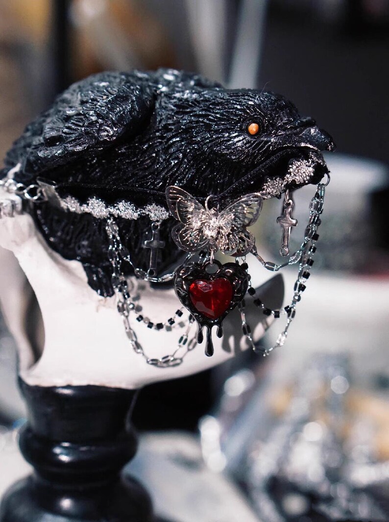 K&ouml;nnte beinhalten: Eine schwarze Rabenfigur mit einem silbernen Schmetterlingsanh&auml;nger, einem roten Herzanh&auml;nger und silbernen Ketten. Der Rabe hat ein rotes Auge und der Herzanh&auml;nger hat eine tropfende rote Fl&uuml;ssigkeit.
