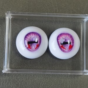 May include: Two purple and white doll eyes with red and black painted details. The eyes are in a clear plastic container.