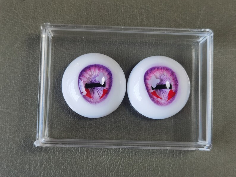 May include: Two purple and white doll eyes with red and black painted details. The eyes are in a clear plastic container.
