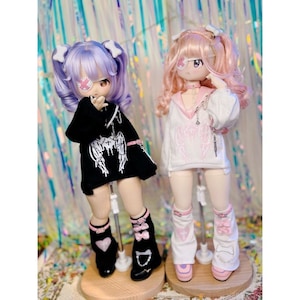 K&ouml;nnte beinhalten: Zwei Puppen mit rosa und lila Haaren, die verschiedene Outfits tragen. Die Puppe links tr&auml;gt ein schwarzes Hoodie mit wei&szlig;en Engelsfl&uuml;geln und einem schwarz-wei&szlig;en Augenpflaster. Die Puppe rechts tr&auml;gt ein wei&szlig;es Hoodie mit rosa Engelsfl&uuml;geln und einem rosa Augenpflaster.