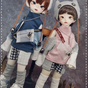May include: Two dolls wearing casual outfits. The doll on the left is wearing a blue and white checkered shirt with a white and gray patterned skirt, a white hat with brown straps, and white and gray socks. The doll on the right is wearing a pink and gray shirt with a black and white checkered skirt, a gray hat with cat ears, and white socks. Both dolls are wearing black sneakers.