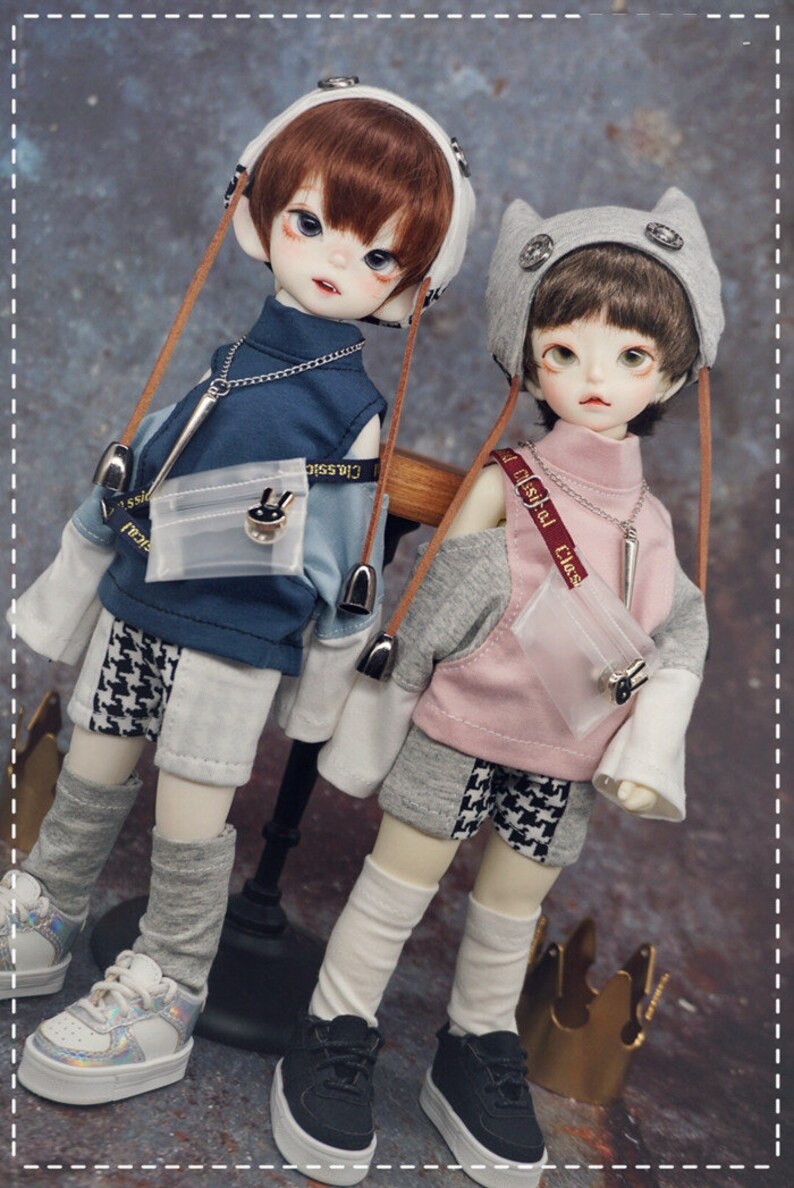 May include: Two dolls wearing casual outfits. The doll on the left is wearing a blue and white checkered shirt with a white and gray patterned skirt, a white hat with brown straps, and white and gray socks. The doll on the right is wearing a pink and gray shirt with a black and white checkered skirt, a gray hat with cat ears, and white socks. Both dolls are wearing black sneakers.