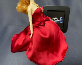 Op de afbeelding: Een rode satijnen baljurk met een sweetheart-halslijn en een grote strik in de taille. De pop draagt een gouden ketting met een rode edelsteen hanger.