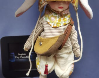 May include: A small doll with a brown and white striped shirt, white pants, and a brown and yellow hat with bunny ears. The doll is wearing a brown leather satchel with a red tassel.