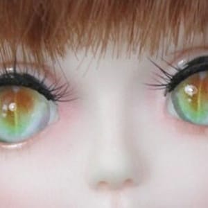 K&ouml;nnte beinhalten: Nahaufnahme eines Puppenkopfes mit gr&uuml;nen und braunen Augen. Die Augen haben ein wirbelndes Muster und sind von langen Wimpern umgeben.
