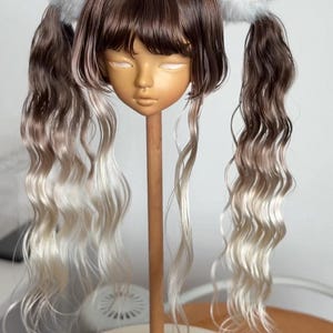 Può includere: Una testa di bambola con capelli castano e biondo ombré acconciati a codini, ognuno adornato con un soffice pompon bianco. La testa della bambola è montata su un supporto di legno, con il numero "18.5" visibile sulla base.