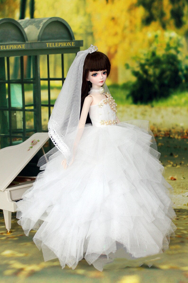 May include: A white bridal gown with a tulle skirt and a lace trim on the veil. The doll is wearing a tiara and a pearl necklace.