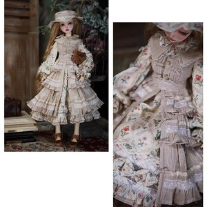 May include: A doll wearing a beige dress with floral print and lace trim. The dress has a ruffled skirt and a matching hat. The doll is holding a small brown teddy bear.