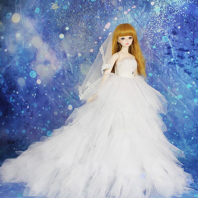 May include: A white tulle wedding dress with a long train and a lace trim. The dress is being worn by a doll with blonde hair and a veil.