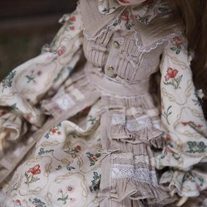 May include: A doll wearing a beige dress with a floral pattern and lace trim. The dress has a ruffled skirt and a matching bodice with a collar.