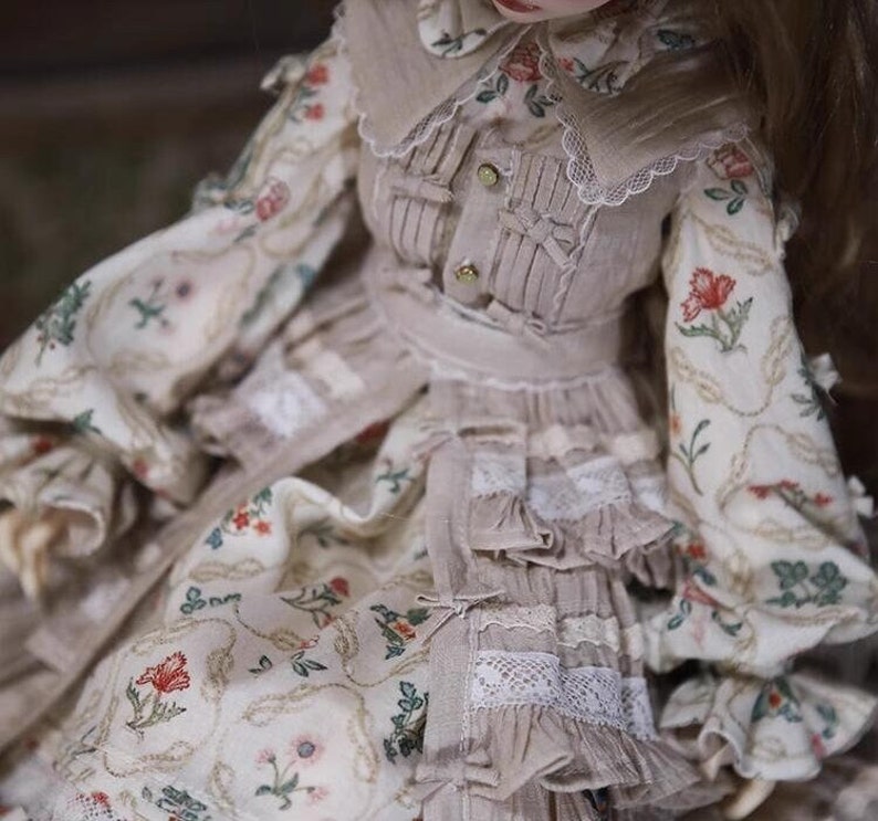 May include: A doll wearing a beige dress with a floral pattern and lace trim. The dress has a ruffled skirt and a matching bodice with a collar.