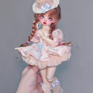 May include: A doll with pink hair in braids, wearing a pink floral dress and a cupcake-shaped hat with a cherry on top. The doll has pale skin, rosy cheeks, and is wearing white shoes.