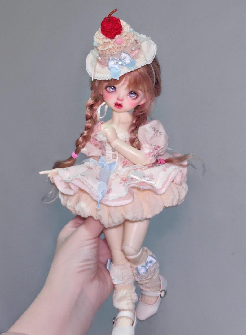 May include: A doll with pink hair in braids, wearing a pink floral dress and a cupcake-shaped hat with a cherry on top. The doll has pale skin, rosy cheeks, and is wearing white shoes.
