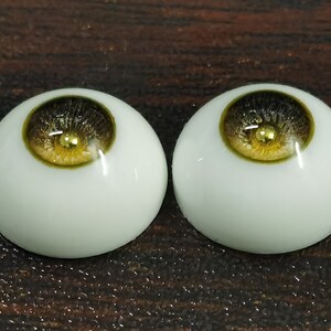 Puede incluir: Dos ojos de mu&ntilde;eca realistas con escler&oacute;tica blanca, iris marr&oacute;n oscuro y una mancha dorada en el centro. El iris tiene un anillo exterior verde.