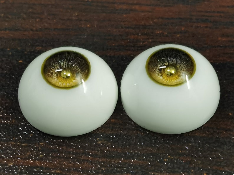 Puede incluir: Dos ojos de mu&ntilde;eca realistas con escler&oacute;tica blanca, iris marr&oacute;n oscuro y una mancha dorada en el centro. El iris tiene un anillo exterior verde.