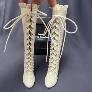 Pode incluir: Um par de botas brancas com atacadores e acabamento ondulado. As botas t&ecirc;m cano alto e s&atilde;o feitas de um material liso. Os atacadores brancos cruzam-se na frente. As botas s&atilde;o para uma boneca.