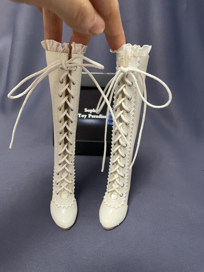 Pode incluir: Um par de botas brancas com atacadores e acabamento ondulado. As botas t&ecirc;m cano alto e s&atilde;o feitas de um material liso. Os atacadores brancos cruzam-se na frente. As botas s&atilde;o para uma boneca.