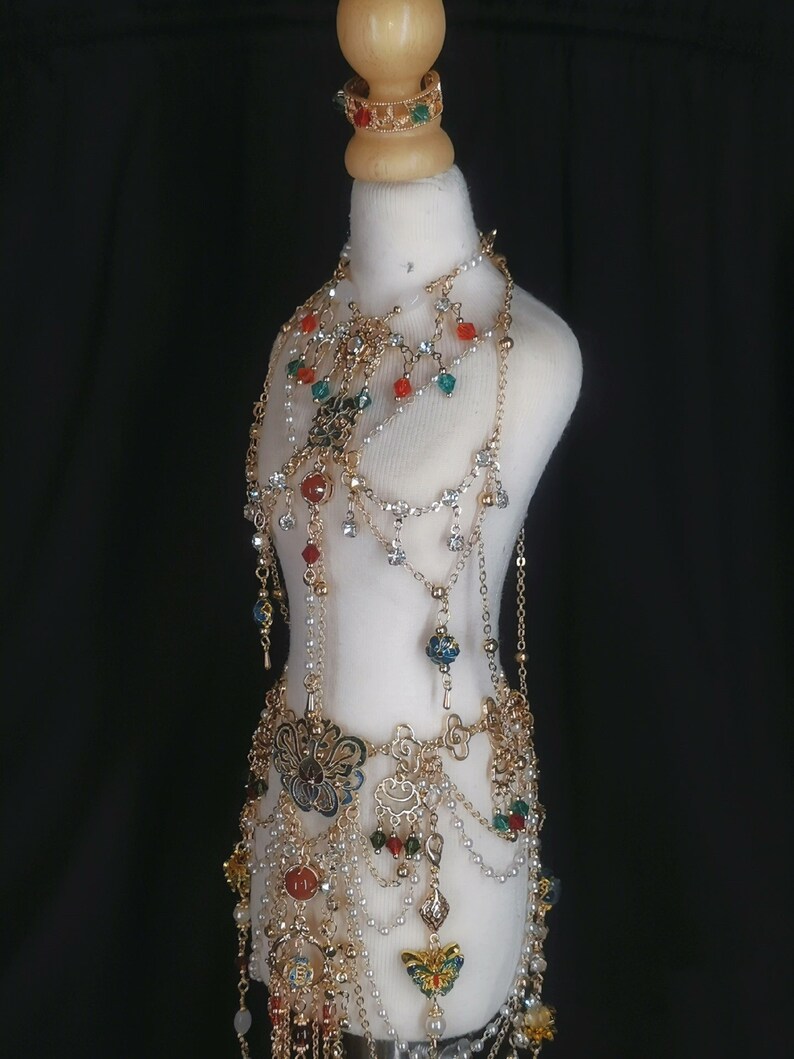 May include: A gold-toned jewelry set with a variety of beads and charms, including flowers, butterflies, and geometric shapes. The set includes a necklace, a belt, and a bracelet. The jewelry is displayed on a white mannequin.