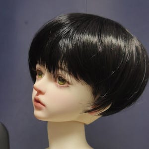 May include: A ball-jointed doll head featuring pale skin, light green eyes, and a short, straight black wig with bangs. The doll has a neutral facial expression with soft pink lips and a defined nose, presented in a profile view against a dark background.