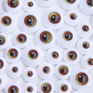 Peut inclure: Assortiment d'yeux de poup&eacute;e bruns et dor&eacute;s de diff&eacute;rentes tailles. Les yeux sont dispos&eacute;s sur un fond blanc.