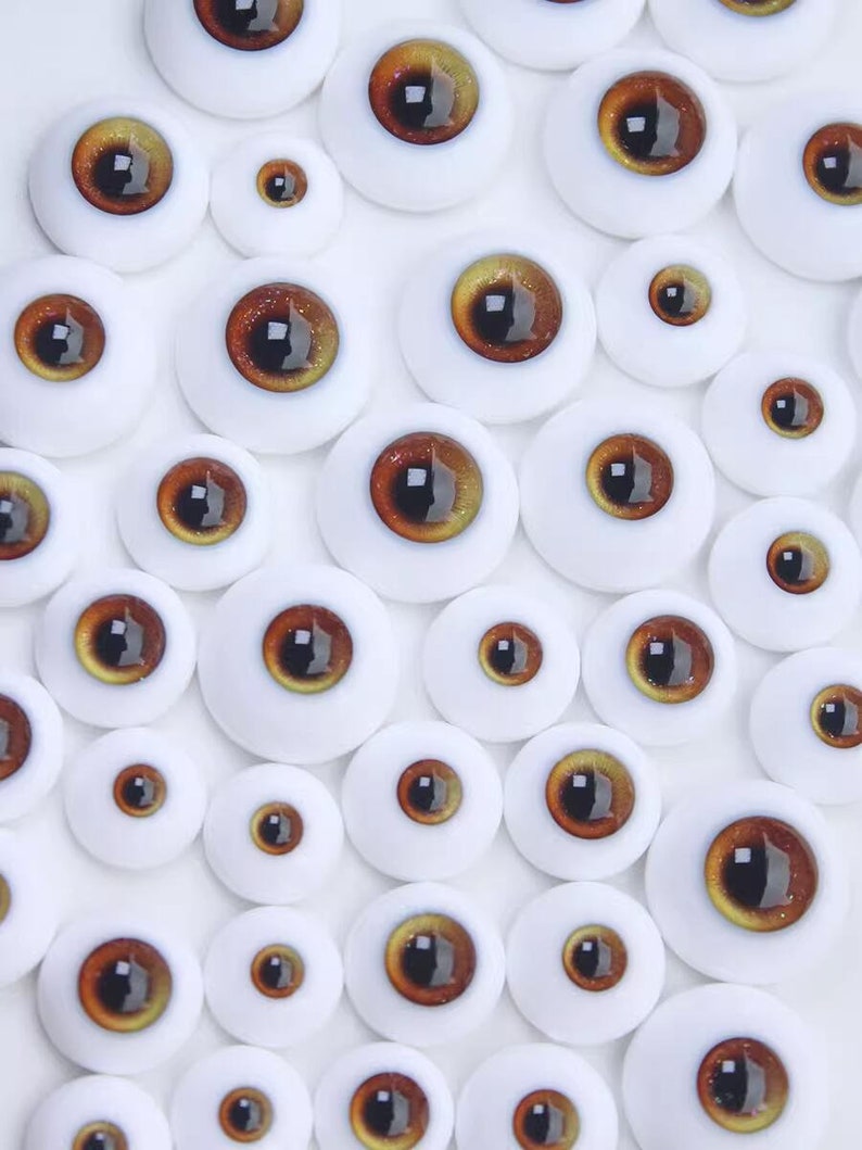 Peut inclure: Assortiment d'yeux de poup&eacute;e bruns et dor&eacute;s de diff&eacute;rentes tailles. Les yeux sont dispos&eacute;s sur un fond blanc.
