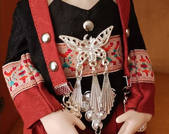 May include: A doll wearing a red and black traditional outfit with silver jewelry. The jewelry features a butterfly design and dangling tassels.