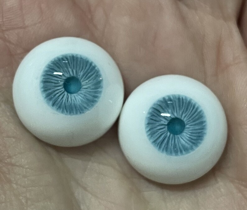 K&ouml;nnte beinhalten: Zwei realistisch aussehende Glasaugen mit wei&szlig;er Sklera und blauen Iris. Die Iris haben ein detailliertes Muster und einen t&uuml;rkisfarbenen Mittelpunkt.