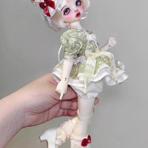 May include: A doll with white hair and blue eyes, wearing a green floral dress, white bloomers, and white shoes with red bows. The doll has a hat with a cherry and cream-colored accents. The doll is approximately 12 inches tall.