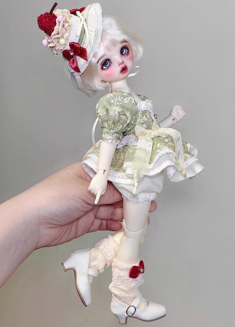 May include: A doll with white hair and blue eyes, wearing a green floral dress, white bloomers, and white shoes with red bows. The doll has a hat with a cherry and cream-colored accents. The doll is approximately 12 inches tall.