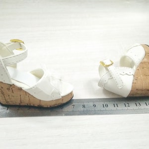 Peut inclure: Une paire de sandales compens&eacute;es blanches avec des talons en li&egrave;ge et des boucles dor&eacute;es. Les sandales mesurent 7 cm de long.
