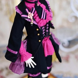Könnte beinhalten: Eine Puppe trägt eine schwarze Jacke mit goldenen Knöpfen und ein rosa Rüschenhemd. Die Jacke hat ein Schnürkorsettdetail und einen rosa Rüschenrock. Die Puppe hat spitze Ohren und trägt schwarze Strumpfhosen.