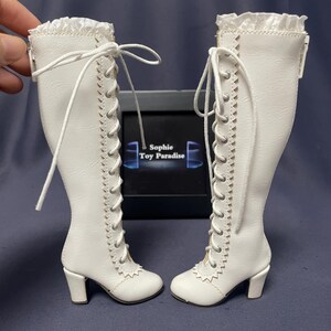 Pode incluir: Um par de botas de boneca brancas com design de cadar&ccedil;o e saltos pequenos. As botas t&ecirc;m uma borda com babados na parte superior e costuras decorativas nas laterais. S&atilde;o exibidas em frente a uma caixa preta com o texto "Sophie Toy Paradise".
