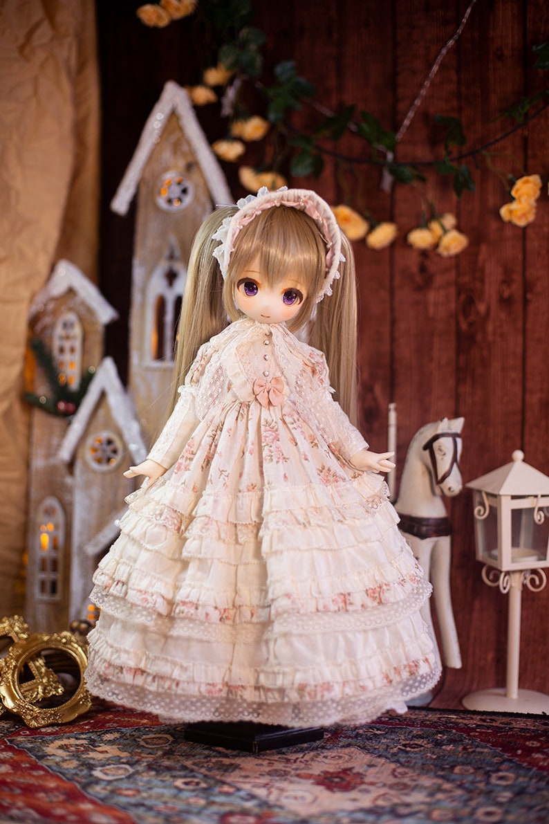 May include: A doll with long blonde pigtails wearing a white dress with pink floral accents and lace trim. The doll is standing on a rug with a wooden house and a white horse in the background.
