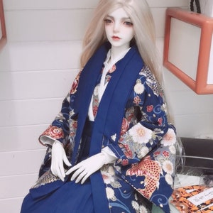 May include: A doll wearing a blue and white kimono with a floral pattern. The kimono has a long, flowing sleeve and a wide collar. The doll has long, white hair and a delicate face.