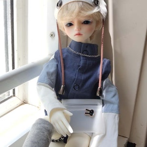 May include: A doll with blonde hair wearing a white hat with bunny ears, a blue shirt, and a white purse with a bunny charm. The doll is sitting on a windowsill.