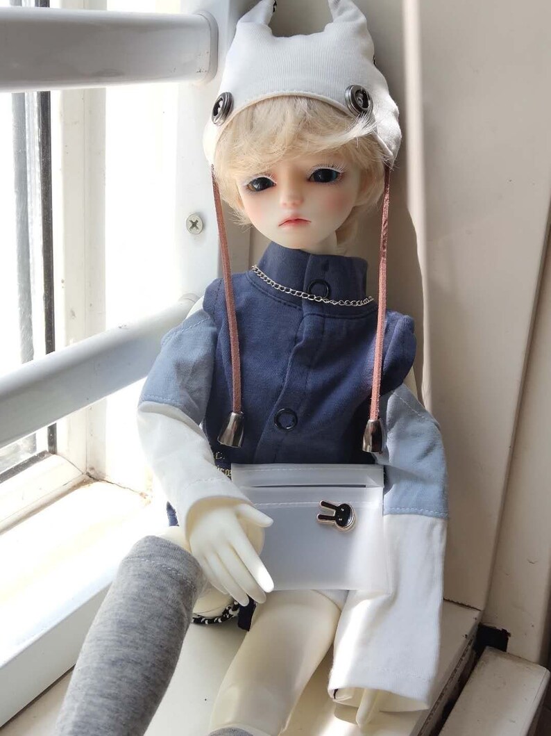 May include: A doll with blonde hair wearing a white hat with bunny ears, a blue shirt, and a white purse with a bunny charm. The doll is sitting on a windowsill.