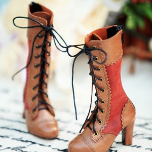 May include: A pair of miniature brown and red lace-up boots with high heels. The boots feature a two-tone design, with brown leather and red suede panels. Black laces and zippers complete the look. The boots are designed for a doll.