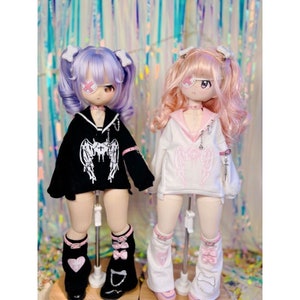 K&ouml;nnte beinhalten: Zwei Puppen mit rosa und lila Haaren, die schwarze und wei&szlig;e Outfits mit Engelsfl&uuml;geln tragen. Die Puppen tragen Augenklappen und haben rosa Schleifen im Haar.