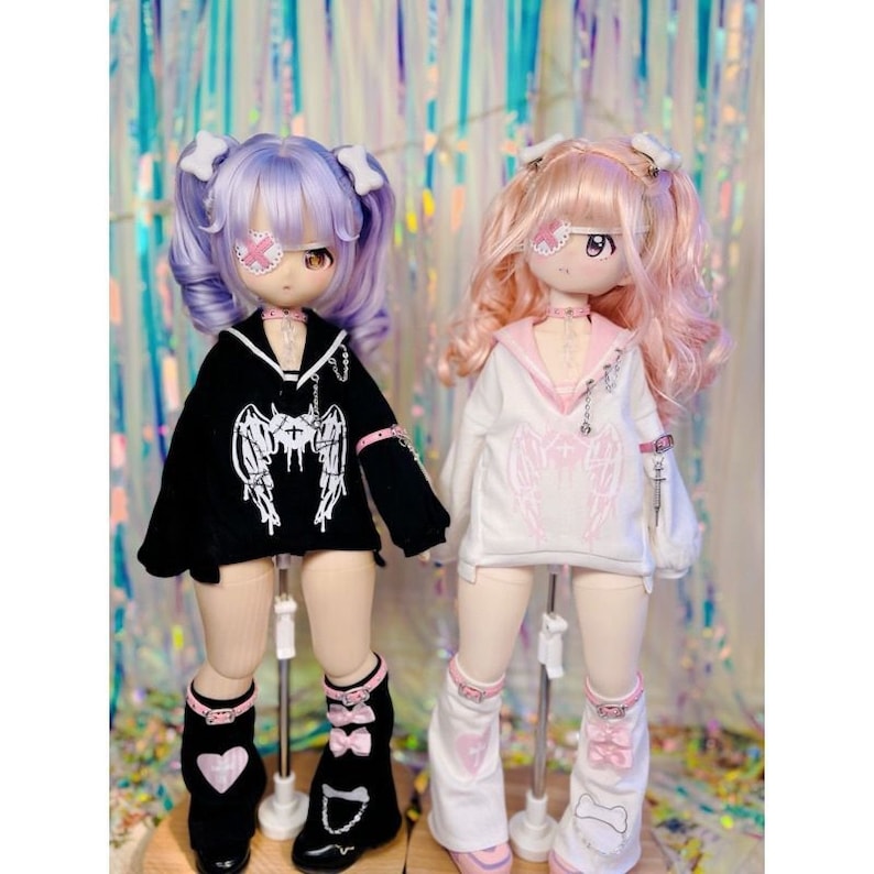 K&ouml;nnte beinhalten: Zwei Puppen mit rosa und lila Haaren, die schwarze und wei&szlig;e Outfits mit Engelsfl&uuml;geln tragen. Die Puppen tragen Augenklappen und haben rosa Schleifen im Haar.