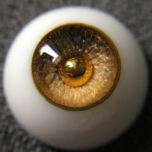 Puede incluir: Primer plano de un ojo de mu&ntilde;eca con un borde dorado y un iris marr&oacute;n y dorado con purpurina.