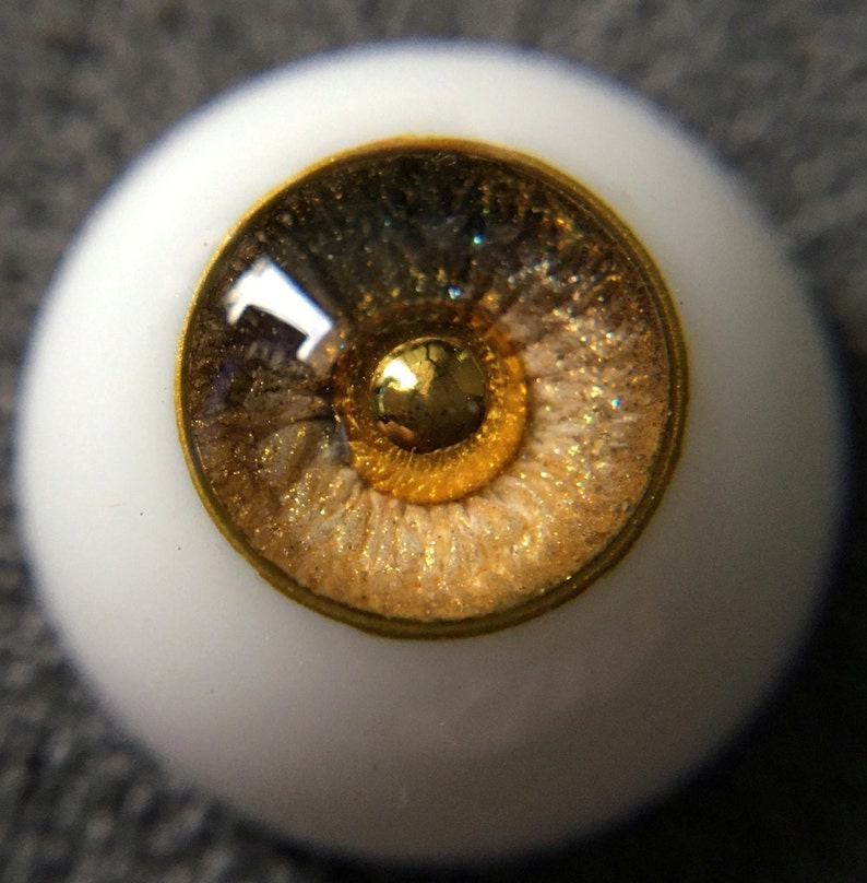 Puede incluir: Primer plano de un ojo de mu&ntilde;eca con un borde dorado y un iris marr&oacute;n y dorado con purpurina.