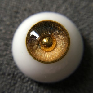 Puede incluir: Primer plano de un ojo de vidrio realista con un iris bordeado de oro y un dise&ntilde;o moteado marr&oacute;n y dorado. El ojo est&aacute; colocado en una carcasa de pl&aacute;stico blanco.
